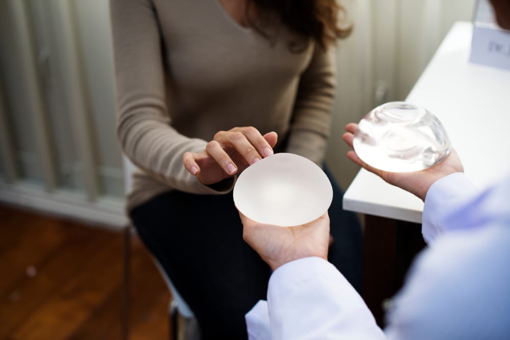 Patient touching a breast implant during consultation, while surgeon hold implant options for hybrid breast reconstruction discussion.
