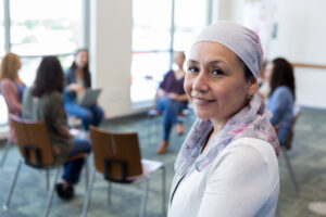 Image of a woman wearing scarf on head while going through cancer treatment, with support group of women sitting in chairs and talking behind her.